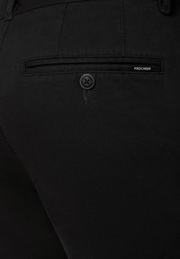 Czarne spodnie o teksturowanej tkaninie, z tylną kieszenią z zapięciem na guzik oraz małą metką "PROCHNIK" nad kieszenią.
