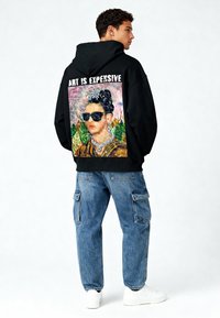 Felpa nera con grafica colorata sul retro di una donna con occhiali da sole. Abbinata a jeans cargo azzurri chiari e sneakers bianche.