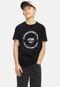 Camiseta de algodón negra con texto blanco en un diseño circular. Combinada con pantalones grises y una gorra de béisbol beige.