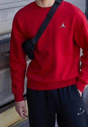 Person trägt rotes Sweatshirt und schwarze Hose mit kleinen Logos, trägt eine schwarze Umhängetasche und steht neben einer Metallwand.