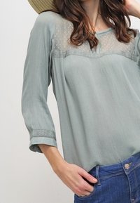 Blusa verde chiaro, con texture e un pannello di pizzo trasparente sul petto, maniche a tre quarti, abbinata a jeans in denim blu.