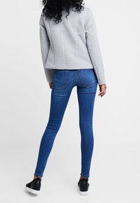 Graue strukturierte Jacke, blaue Skinny-Jeans und schwarze Sneakers, von hinten betrachtet, um das taillierte Design und die Denim-Wäsche hervorzuheben.