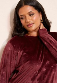 Top lung cu maneci lungi din catifea crimson, cu texturăîn relief și un model subtil de stele. Design cu guler rotund, maneci ajustate și finisaj ușor lucios.