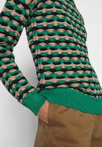 Pull tricot texturé vert, noir et beige porté avec un pantalon marron, main dans la poche, sur un fond uni.