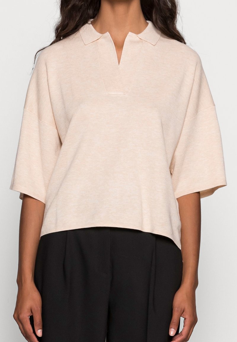 Femme portant un top en maille beige ample à manches courtes avec un col fendu, associé à un pantalon noir taille haute, vue du cou aux hanches.
