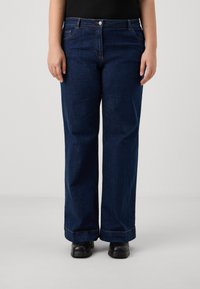 Jean en denim bleu foncé, coupe ample, avec des détails de couture subtils, cinq poches et une taille mi-haute, associés à des chaussures noires.