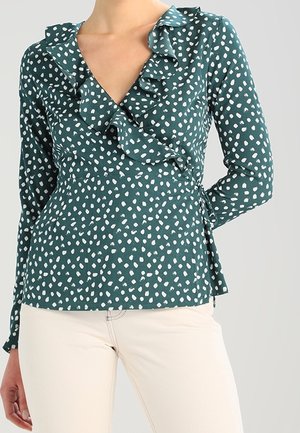 Groene wikkelblouse met witte onregelmatige stippen, geruffleerde halslijn, lange mouwen, knoop in de taille, gedragen met lichtbeige broek.