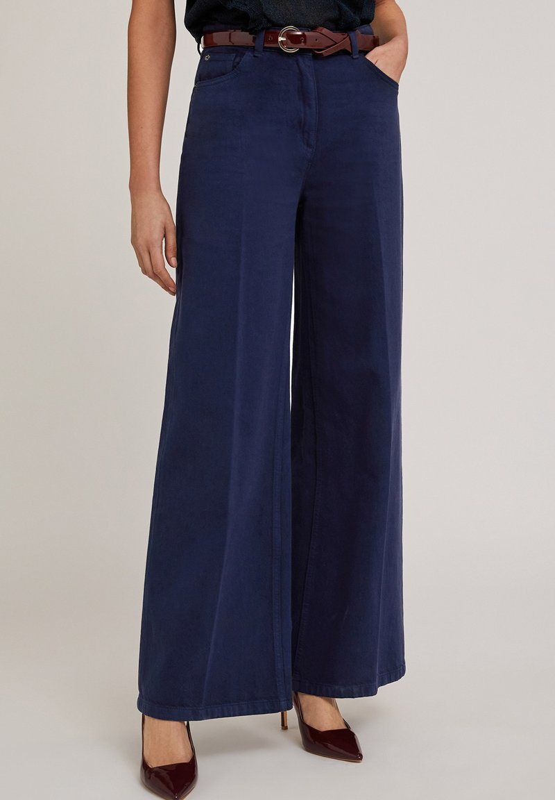 Hoge taille, wijde marineblauwe pantalons gemaakt van gladde stof. Heeft voorzakken en een bruine riem met een zilveren ronde gesp.