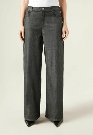 Czarne spodnie o szerokich nogawkach z zapięciem na guzik i zamek błyskawiczny, noszone z czarnymi, spiczastymi butami oraz białym topem widocznym w talii.