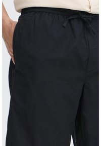 Black cotton trousers with an elastic waistband, drawstring detail, and side pockets. Smooth texture and relaxed fit.