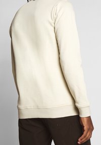 Kremowa bluza z długimi rękawami i prążkowanymi mankietami. Gładka faktura i luźny krój, zestawiona z ciemnobrązowymi spodniami.