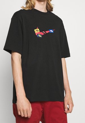 Osoba nosząca czarną koszulkę z czerwonym logo Nike i trzema naszywkami z logo Jumpman w kolorze żółtym, niebieskim i czarnym na piersi.