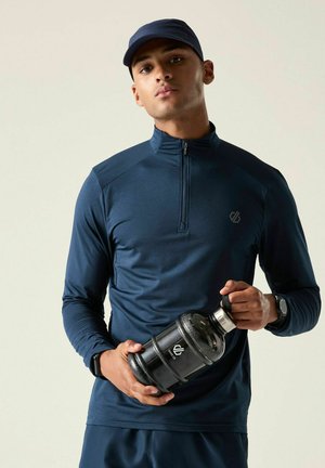Man in navy sportswear and cap holding a large black water bottle with a protective sleeve, standing against a plain background.