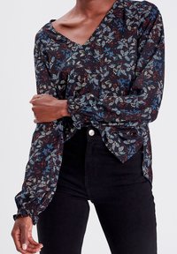 Chemisier floral en tissu sombre avec des motifs bleus et rouges, décolleté en V et poignets élastiqués. Associé à un jean noir.