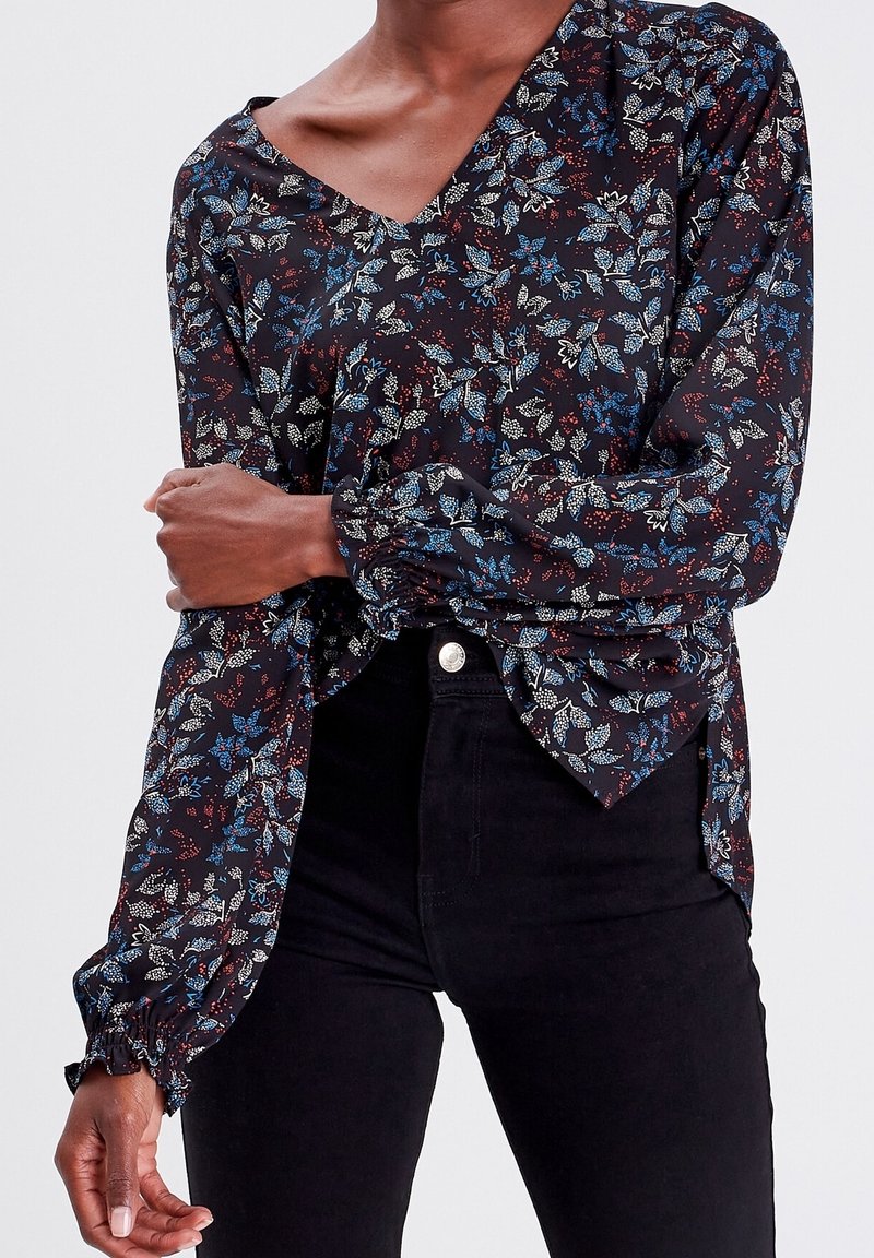 Chemisier floral en tissu sombre avec des motifs bleus et rouges, décolleté en V et poignets élastiqués. Associé à un jean noir.