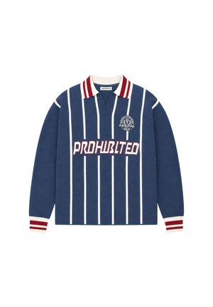 Homme aux cheveux courts portant un pull rayé bleu marine avec un col et des poignets rouges et blancs, tenant un tissu rouge, regardant de côté.