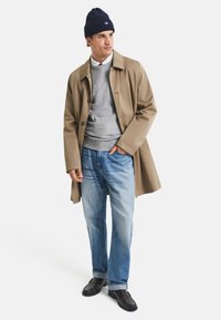 Cappotto di cotone beige sopra un maglione grigio, jeans di denim blu e mocassini neri. Il cappello è blu navy con un logo. Il modello sta in piedi con le mani in tasca.