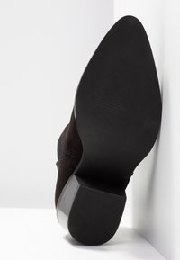 Bottine en daim noir posée avec la semelle contre un mur blanc, montrant une semelle extérieure pointue, lisse et légèrement texturée ainsi qu'un talon bloc.