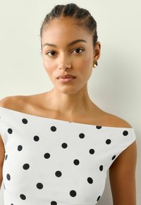 Biała bluzka bez ramiączek w czarne kropki. Gładki materiał, dopasowany krój, szeroki dekolt oraz minimalistyczne złote kolczyki.