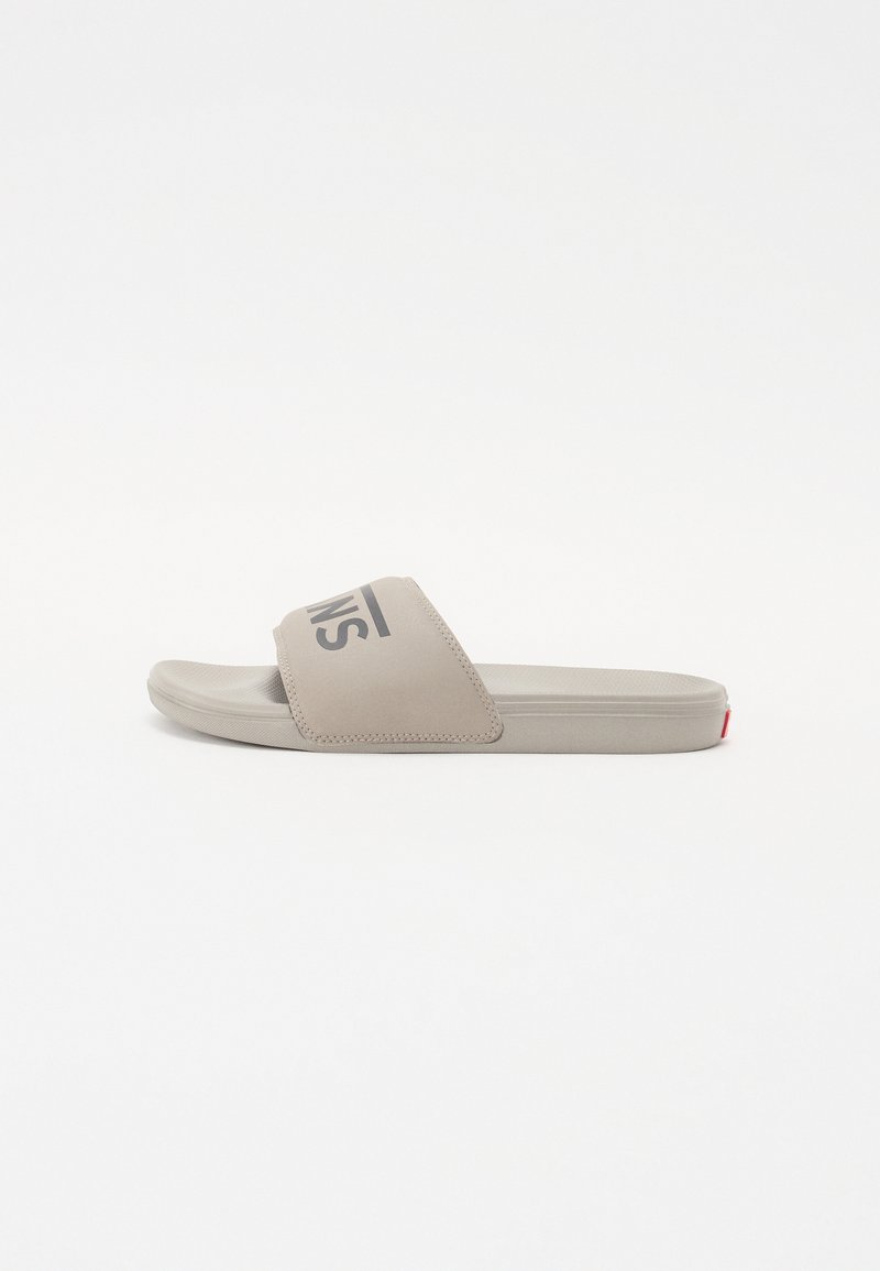 Sandale à semelle plate en beige clair, avec une large sangle ornée d'un logo noir, en matériau texturé lisse, et une subtile accent rouge au niveau du talon.