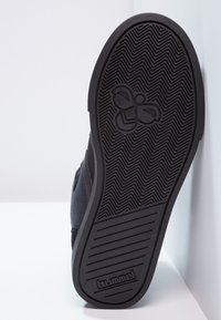 Sneeaker preto com uma sola de borracha texturizada exibindo um padrão em zig-zag e um logótipo. A parte superior apresenta uma mistura de materiais, com acabamentos costurados.