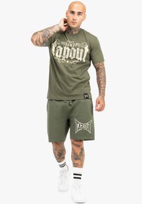 Maslinasto zeleni pamučni majica s velikim bijelim natpisom "Tapout" i odgovarajućim kratkim hlačama. Zapaženi logo detalj i kontrastne bijele tenisice s prugama.