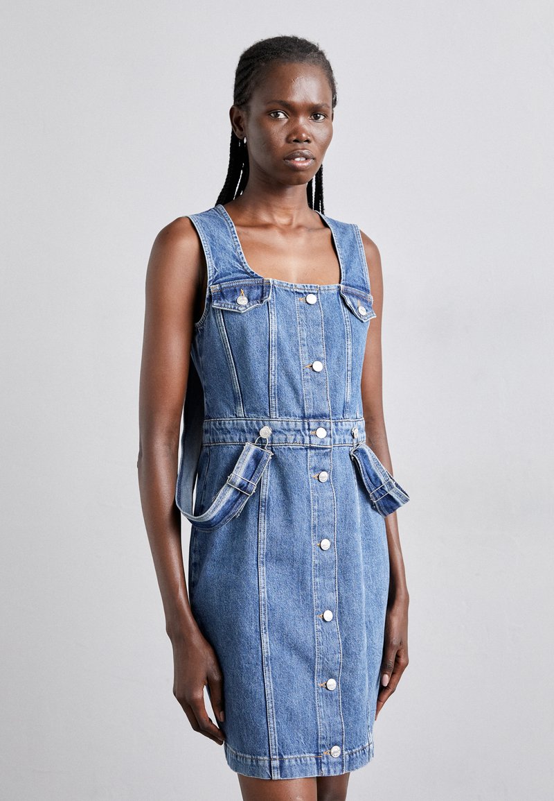 Denim kleita ar kvadrātveida kakla izgriezumu, bez piedurknēm, priekšējām pogām un divām atloku kabatām. Gaiši zila krāsa ar gludu tekstūru.