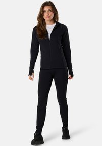 Svart zip-up jacka med lång ärm, i kombination med matchande tajta byxor. Slät textur, sportig design och praktiska fickor med dragkedja.