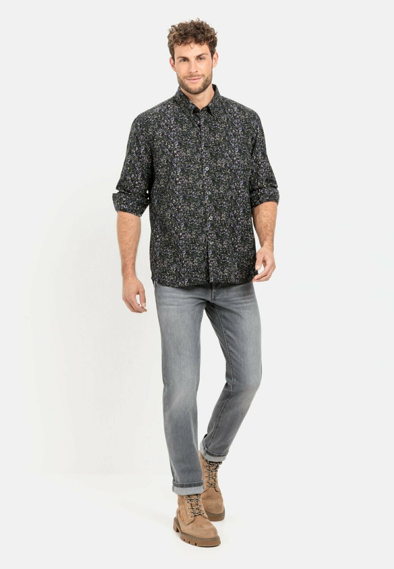 Schwarzes Hemd mit Knöpfen und floralem Muster, graue Jeans und beige Schnürstiefel. Das Hemd hat lange Ärmel, die bis zu den Ellbogen hochgerollt sind.