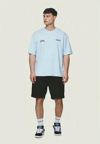 Lichtblauw katoen T-shirt met korte mouwen, met zwarte grafische tekst. Gecombineerd met zwarte shorts, witte sokken en marineblauwe sneakers.