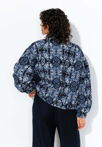 Veste à motif marine avec des manches ballon, présentant un design géométrique dans des teintes bleues et blanches. Tissu doux avec une coupe décontractée.