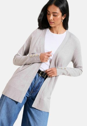 Žena v svetlosivej otvorenej kardigán so zlatými gombíkmi na manžetách, bielom topu, modrých džínsoch s vysokým pásom a čiernym opaskom, pozerajúca sa dole.