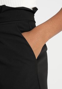 Tessuto di pantaloni neri con una texture liscia; presenta una tasca laterale con una mano parzialmente infilata all'interno; la vita è strutturata e piegata.