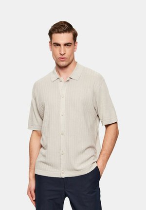 Homme portant une chemise en maille beige clair à manches courtes avec des rayures verticales subtiles et un pantalon foncé, debout avec une main dans la poche.