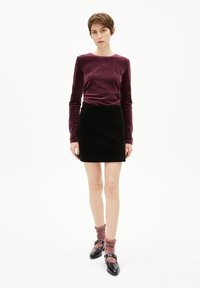 Burgunderfarbene Samtoberteil mit langen Ärmeln, schwarzer Cord-Minirock, rosa und grau gestreifte Socken und schwarze Schuhe mit Schnalle.