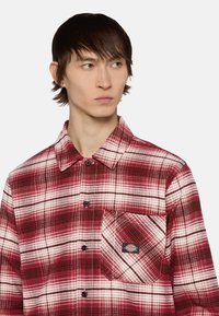 Rood-wit geruite flanellen shirt met een knoopsluitingen, een borstzak en een logo patch. De stof lijkt textuur te hebben en is warm.