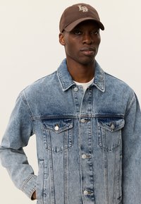 Τζιν μπουφάν σε ανοιχτό μπλε χρώμα με ξεθωριασμένη υφή, διαθέτοντας μπροστινά κουμπιά, δύο τσέπες στο στήθος και κλασικό γιακά. Καφέ καπέλο με λογότυπο.