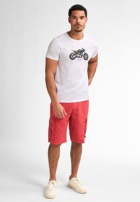 Camiseta blanca con un gráfico de motocicleta, combinada con pantalones cortos rojos de cargo con bolsillos laterales. El modelo lleva zapatillas color crema.