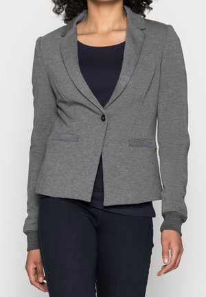 Mujer con un blazer ajustado gris sobre una blusa azul marino oscuro y pantalones oscuros, de pie contra un fondo claro y liso.