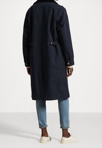 Abrigo largo azul marino con un corte relajado, que cuenta con un collar de tela de pana, detalles de botones y bolsillos laterales, combinado con jeans azul claro y zapatillas deportivas.