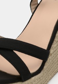 Sandale compensée beige avec semelle plateforme en corde tressée et brides croisées en tissu noir sur le pied.
