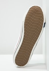 Sneaker mit einem weißen Gummi-Obermaterial, brauner strukturiertem Außensohle mit Wellenmustern und dezenten roten und blauen Streifen an den Seiten.