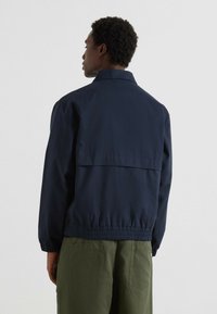 Giubbotto anti-vento della marina con elastico in vita, spalle scese e dettaglio di apertura sul retro. Abbinato a pantaloni cargo verdi.