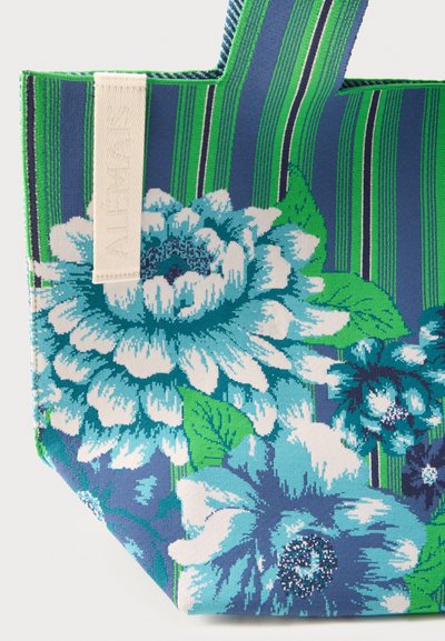 Τσάντα tote με μπλε και πράσινες ρίγες, μεγάλα μπλε και λευκά λουλουδένια σχέδια, πράσινα φύλλα και μια μπεζ υφασμάτινη ετικέτα πάνω αριστερά.
