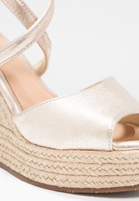 Sandale compensée avec empeigne à bout ouvert métallique doré et semelle plateforme en jute tressé, dotée de brides croisées à la cheville, sur fond blanc.