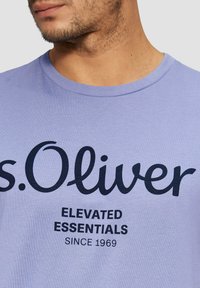 Violetti puuvillapaita, jossa on pyöreä pääntie, ja jossa on suuri tummansinisestä painatuksesta koostuva teksti "s.Oliver" ja "ELEVATED ESSENTIALS".