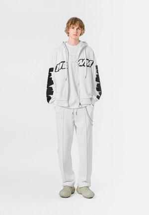 Sudadera blanca con detalles negros y logo en las mangas, combinada con pantalones de chándal blancos. Calzado gris claro con un diseño texturizado.