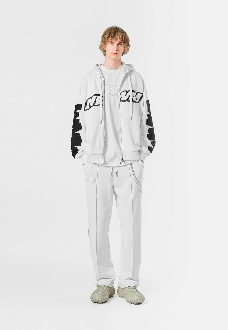 Sudadera blanca con detalles negros y logo en las mangas, combinada con pantalones de chándal blancos. Calzado gris claro con un diseño texturizado.