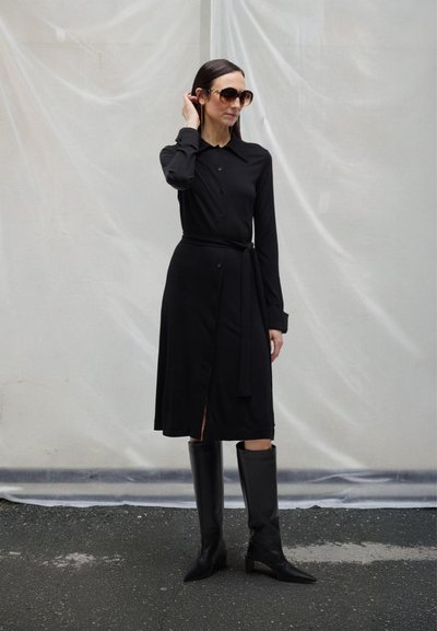 Vrouw die een zwart, lang mouwig, geknoopt jurk met riem draagt, zwarte laarzen tot aan de knie en grote zonnebril, staand op asfalt tegen een lichte achtergrond.