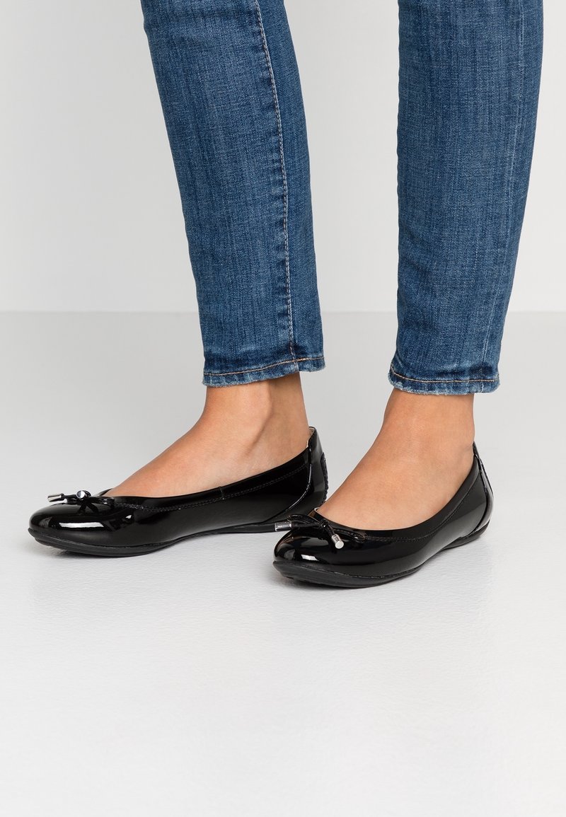 Des ballerines en cuir verni noir avec un bout arrondi, un nœud décoratif à l'avant et une finition lisse et brillante. Portées avec un jean bleu.